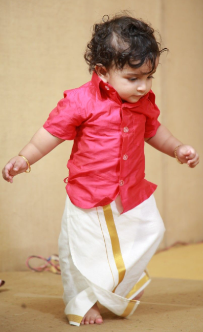 Child wearing a bright pink shirt and Velcro Onam dhoti with gold border on a beige background